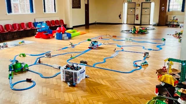 Bright hall with wooden flooring, toy train tracks, colorful playsets, and seating along the walls.