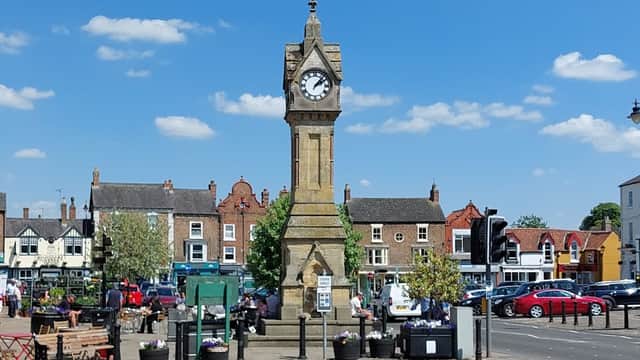 Historic clock tower in a bustling market square, surrounded by shops, greenery, and parked cars under a clear blue sky.