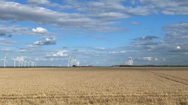 Rolling golden fields under a blue sky with scattered clouds, featuring wind turbines in the distance.