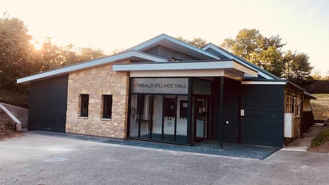 Modern village hall with stone and wooden exterior, large glass entrance, surrounded by greenery and trees.