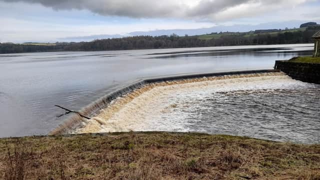Water cascading over a stone dam into a calm reservoir, surrounded by trees and rolling hills under a cloudy sky.