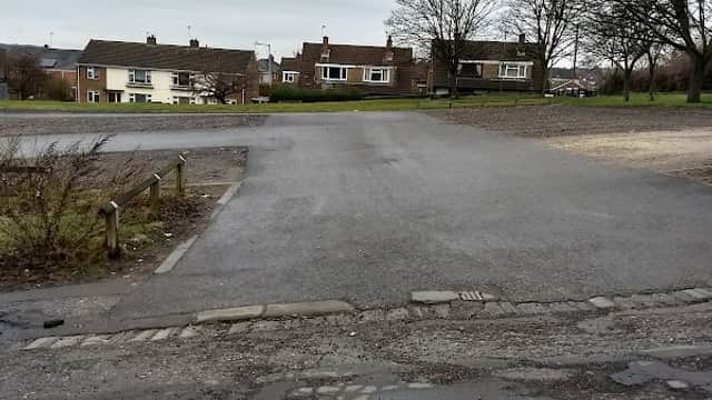 A gravel car park with a few trees, houses in the background, and a cloudy sky overhead.