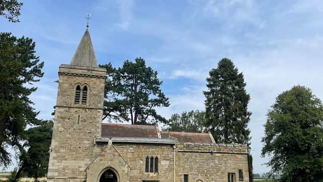 Historic stone church with a tall steeple, surrounded by trees and a grassy graveyard under a blue sky.