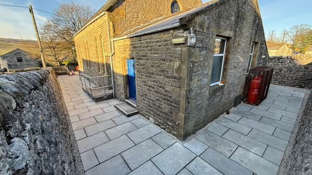 Stone building with a blue door, large windows, and a paved pathway, surrounded by a dry stone wall and trees.