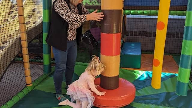 A child in a tutu plays with a colorful padded post while an adult assists in a vibrant indoor play area.