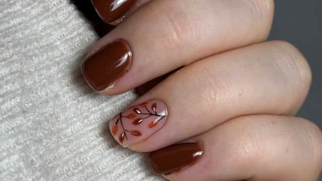 Brown manicure with a leaf design on one nail, hand resting on a light-colored sweater.