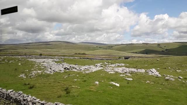 Rolling green hills under a blue sky with fluffy clouds, featuring dry stone walls and scattered rocks in the landscape.