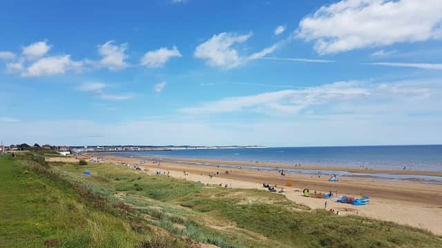 Wide sandy beach with people enjoying the sun, green grass in the foreground, and a clear blue sky with clouds.