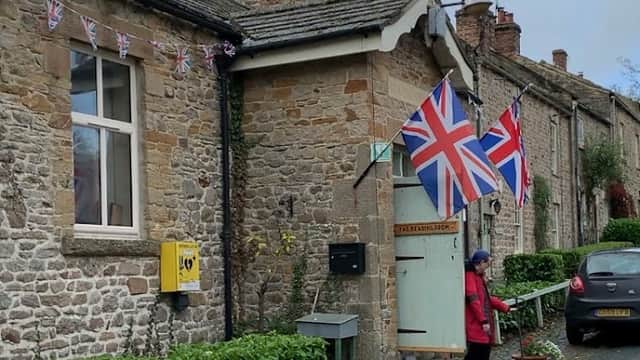 Stone building with Union Jack flags, a woman standing outside, and a parked car in a quaint Yorkshire village.