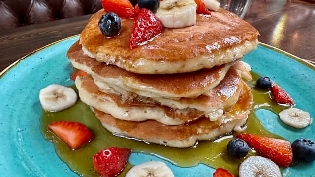 Stack of fluffy pancakes topped with strawberries, blueberries, and banana slices, drizzled with syrup on a teal plate.