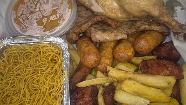 Takeaway box featuring fried chicken, sausages, chips, and noodles, with a container of sauce on the side.
