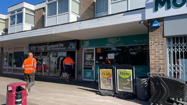 Brightly lit car park with shops, including a sandwich bar, and two workers in high-visibility vests.