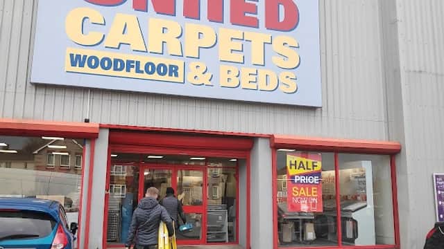 Storefront of United Carpets and Beds with a large sign, a person carrying a ladder, and a parked car.