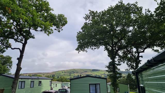 Green caravan units surrounded by trees, with rolling hills in the background under a cloudy sky.