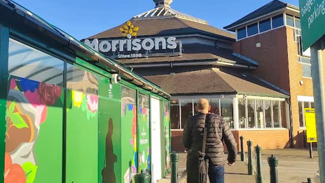 A person walks towards Morrison's Supermarket, with colorful floral murals and clear blue skies in the background.