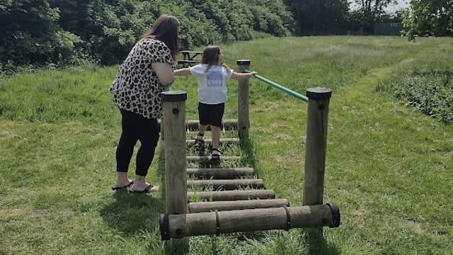 A woman and a child navigate a wooden balance beam in a grassy field surrounded by trees under a cloudy sky.