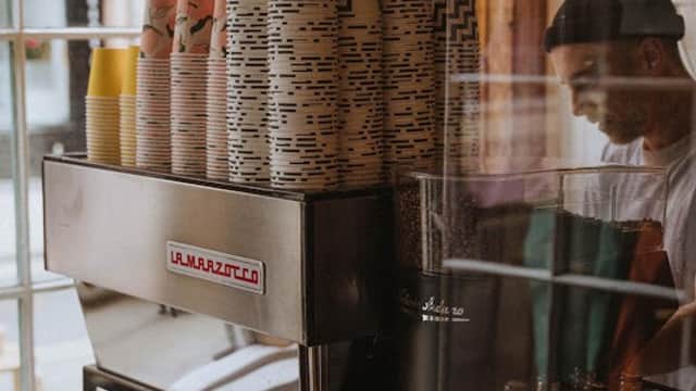 Kiosk Cafe interior featuring a La Marzocco coffee machine, stacked cups, and colorful beverage cans.