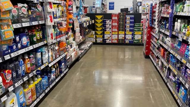 Aisle in Tesco Express with shelves stocked with various household products and groceries, bright lighting overhead.