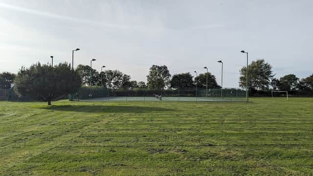 Tennis courts surrounded by neatly trimmed grass and trees, with goalposts in the background under a clear sky.