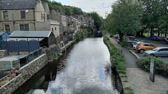 View of a canal beside Station Road car park, with stone buildings, greenery, and parked cars along the pathway.