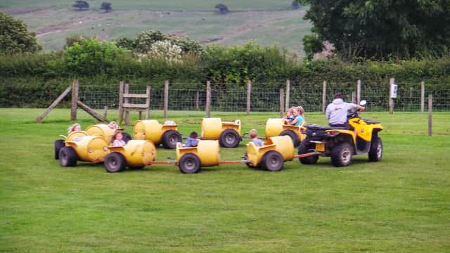 Children enjoying a ride on yellow barrel carts pulled by an ATV in a grassy field surrounded by trees and hills.