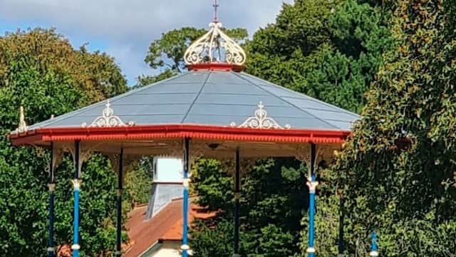 Victorian bandstand surrounded by trees and colorful flower beds under a blue sky with fluffy clouds.