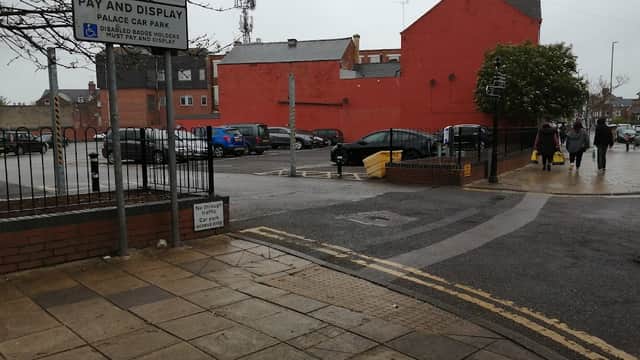 Pay & Display sign at Palace Car Park, with parked cars, a red building, and pedestrians walking in rainy weather.
