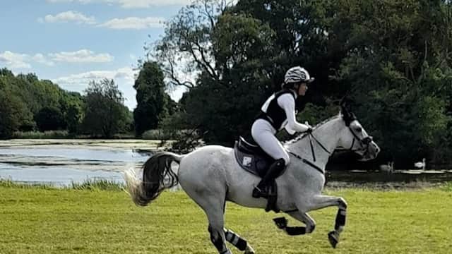 A rider in white attire gallops on a gray horse across a grassy field near a lake and trees under a blue sky.