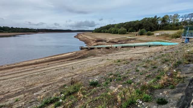 Eccup Reservoir with a dry shoreline, green pipe, and trees lining the water's edge under a cloudy sky.