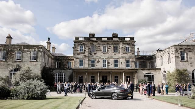 Elegant mansion with guests gathered outside, lush lawn, and a sleek car parked in front under a cloudy sky.