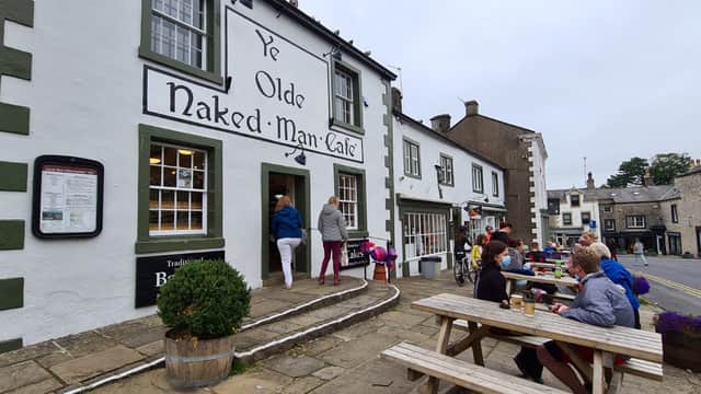 Historic building with "Ye Olde Naked Man CafΓ©" sign, outdoor seating, and people enjoying drinks in Settle, Yorkshire.