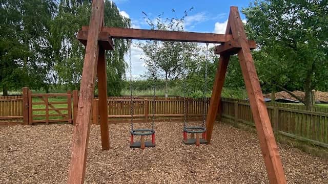 Wooden swing set with two swings, surrounded by a fenced area and lush greenery under a blue sky.