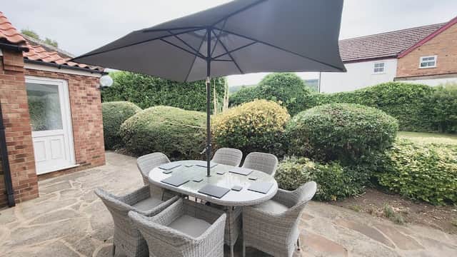 Outdoor seating area with a round table and eight wicker chairs under a large umbrella, surrounded by greenery.