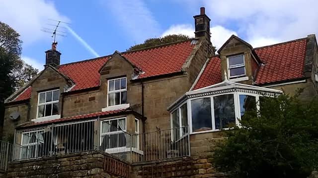 Stone house with red-tiled roof, large windows, and a garden, set against a blue sky with wispy clouds.