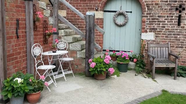 Charming outdoor seating area with white furniture, potted flowers, and a rustic door against a brick wall.