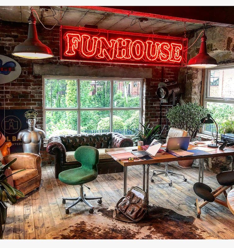 Vintage interior with a neon "FUNHOUSE" sign, leather sofa, green chair, and a desk with a laptop and plants.