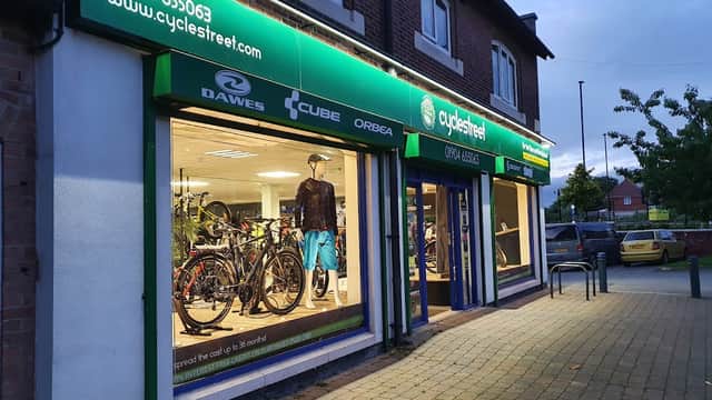Bike shop exterior with illuminated display showcasing bicycles and cycling gear, green signage, and nearby parked cars.