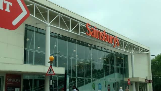 Sainsbury's storefront with large glass windows, red signage, and a few people outside under a cloudy sky.