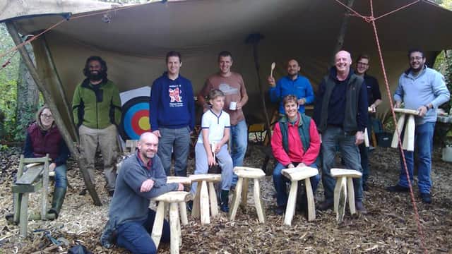 Group of people posing under a large tarp with wooden stools and an archery target in a woodland setting.