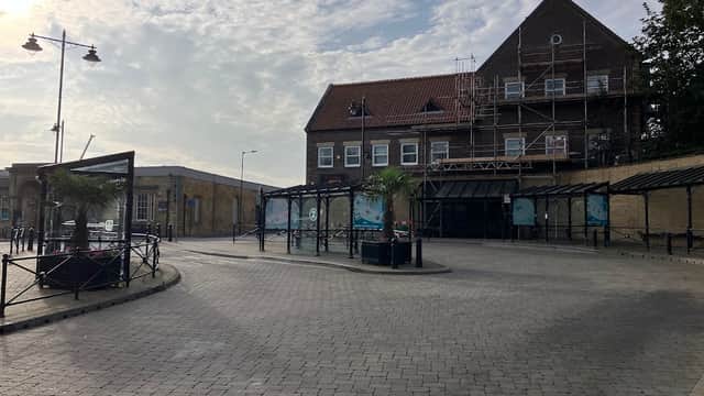 Whitby bus station with a cobblestone area, palm trees, and a building under scaffolding against a cloudy sky.