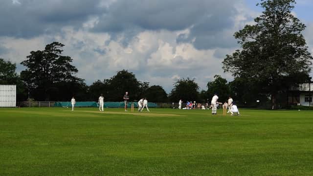 Cricket match in progress on a lush green field, with players in white uniforms and a cloudy sky overhead.