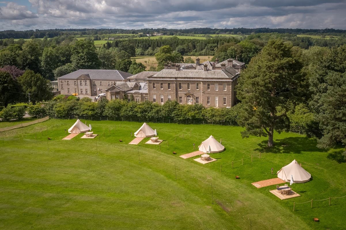 Gisburne Park Estate features a large historic building and glamping tents on a lush green lawn surrounded by trees.