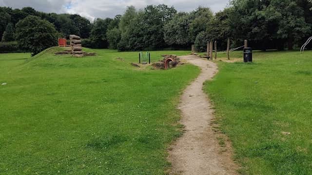 Pathway through a grassy area with trees, play equipment, and a cloudy sky in Kendal Park, Anston.