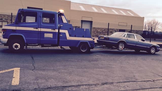 A blue tow truck pulling a silver car on a trailer in a parking lot, with a building in the background.