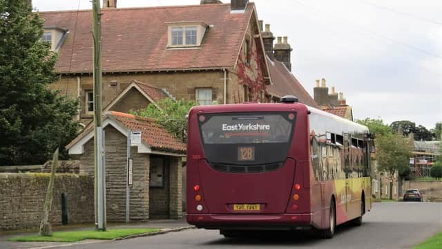 A bus labeled "East Yorkshire 128" drives past a stone building with a red roof in Hutton Buscel, Yorkshire.