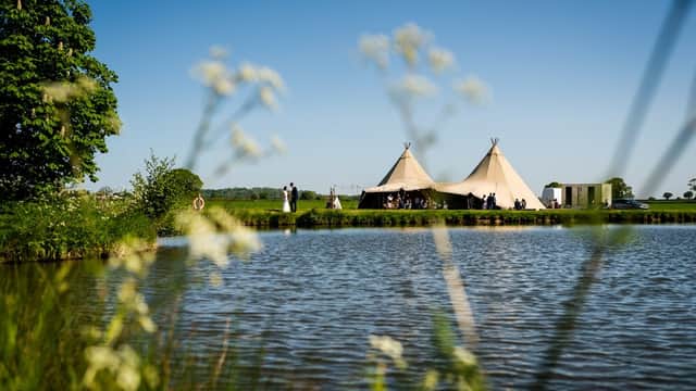 Tipi tents by a serene lake, surrounded by lush greenery and clear blue skies, perfect for a wedding venue.
