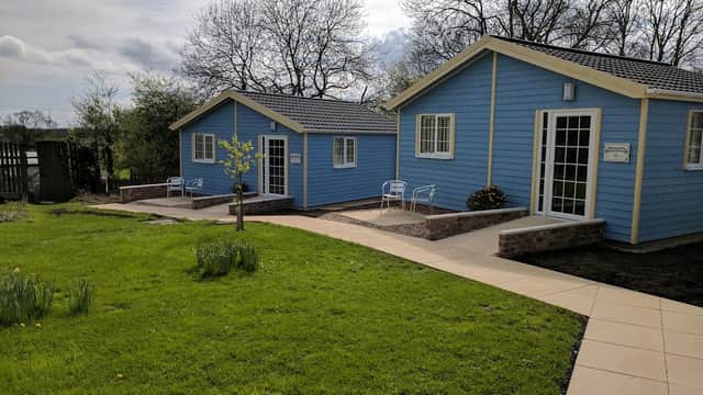 Two blue wooden cottages with white trim, surrounded by green grass and trees, featuring small patios and a pathway.