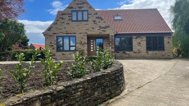 Modern stone house with a red tiled roof, landscaped garden, and a paved driveway under a blue sky.