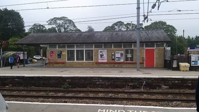 Guiseley Station with a stone building, posters, and a "Mind the Step" warning on the platform. Trees in the background.