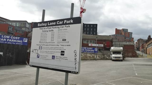 Bailey Lane Car Park sign with parking tariffs, empty lot, and buildings in the background under a cloudy sky.